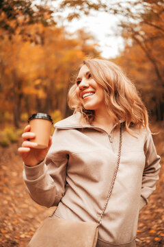 Smiling woman with coffee cup in autumn forest. Joyful tourist in beige sweater enjoying hot drink among yellow trees. Coffee break for nature lover outdoors.
