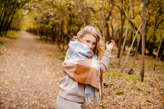 Fashion woman in cozy plaid blanket in autumn forest. Woman traveler wrapped in scarf looking at camera among yellow trees. Concept of keeping warm outdoors.