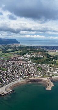 High altitude aerial footage over Penrhyn Bay North Wales looking across the coastal neighbourhood shoreline green countryside distant mountains and expansive sea beneath dramatic clouds