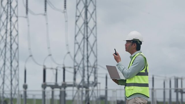 Engineer Conducting Electrical Substation Inspection of Power Equipment Outdoors With Radio and Laptop, Wearing Protective Clothing, Reflective Safety Vest, and Hard Hat During Utility Maintenance
