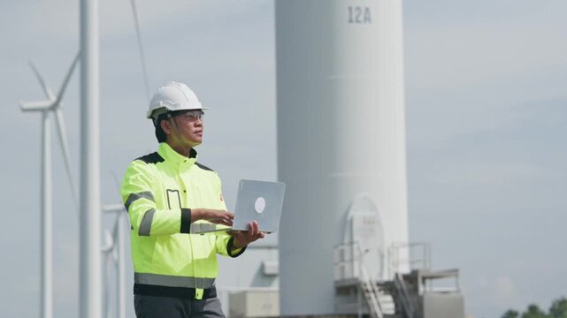 Wind turbine engineer inspecting renewable energy tower with laptop, wearing safety helmet and reflective jacket during outdoor maintenance, sustainable power infrastructure fieldwork and turbine