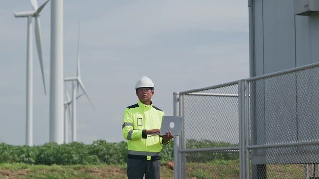 Engineer inspecting wind turbine equipment with laptop, safety helmet, and reflective clothing renewable energy facility near industrial building, advancing clean technology cloudy daylight industry