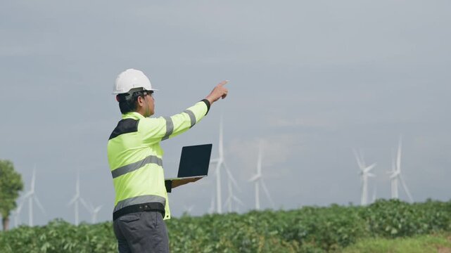 Engineer safety helmet laptop wind turbine field renewable energy inspection, agricultural worker high visibility jacket pointing across landscape during outdoor turbine maintenance assessment cloudy