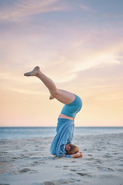 On beach sand, a woman holds Salamba Sirsasana Konasana yoga, a Wide-Legged Headstand, with her head and forearms grounded and raised legs spread apart