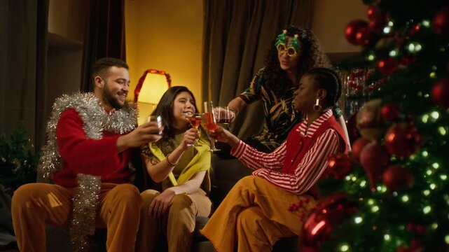 Diverse young adults celebrating winter holidays, toasting drinks on sofa near Christmas tree. Useful for festive season marketing campaigns, beverage commercials and party promotions