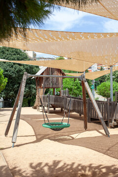 Basket swing and wooden playground equipment located in a public city park under shade sails surrounded by green trees. Outdoor recreational space designed for children, families and active leisure