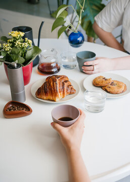hand holding coffee cup. Morning Coffee for Two with Croissants and Chocolate Cookies on a Bright Table with Hand Grinder, Coffee Beans, Kettle and Green Plants. vertical composition