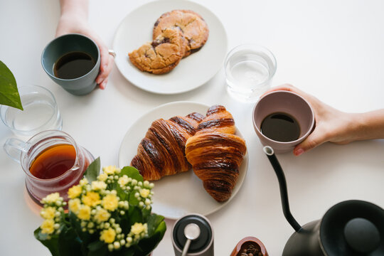 Friends Having Black Coffee and Sharing Chocolate Chip Cookies at a White Breakfast Table with Croissants, Water, Coffee Beans, Hand Grinder, Gooseneck Kettle and Yellow Flowers. top view