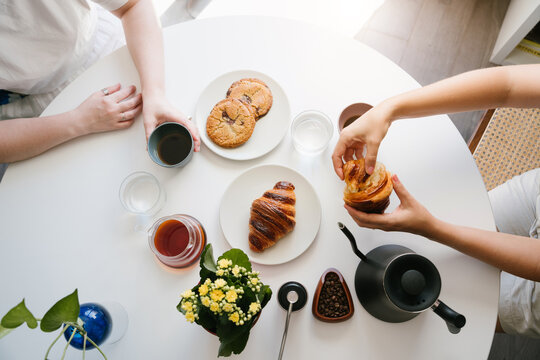 Two Friends Sharing Fresh Pour Over Coffee at a White Table with Croissant, Chocolate Chip Cookies, Water Glasses, Coffee Beans, Hand Grinder, Gooseneck Kettle and Yellow Flowers. top view