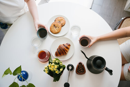 Two Friends Sharing Fresh Pour Over Coffee at a White Table with Croissant, Chocolate Chip Cookies, Water Glasses, Coffee Beans, Hand Grinder, Gooseneck Kettle and Yellow Flowers