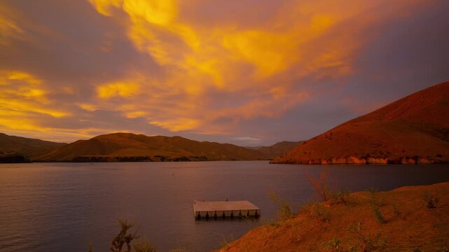 View of Lucky Peak Lake at sunset with calm water and rolling hills under a vibrant golden sky in Ada County, Idaho, United States.