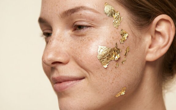 Smiling woman with natural freckles showing luxury 24k gold foil facial mask treatment for skin brightening.