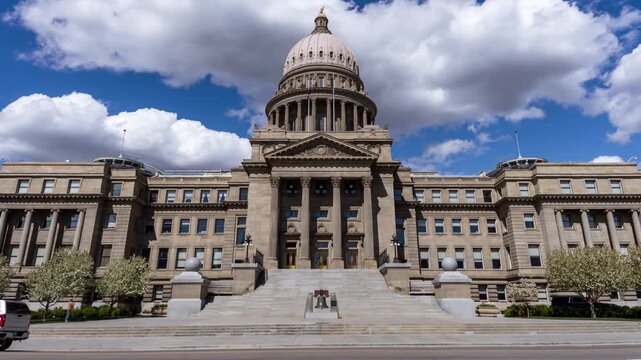 View of the Idaho State Capitol building with its neoclassical dome and stone facade, featuring a white SUV parked in front under a blue sky in Boise, Idaho, United States.
