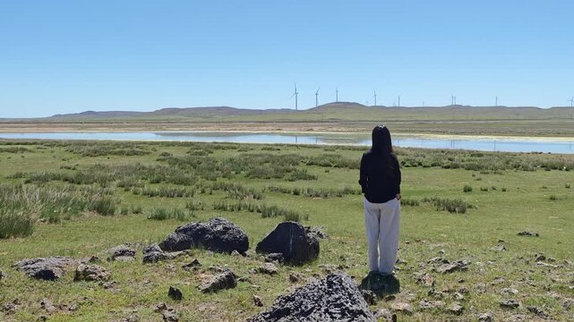 One Asian woman enjoying scenic landscape of salt lake wetland and distant wind turbines in summer in Ulan Hada Volcano Geopark, Ulanqab, Inner Mongolia, China