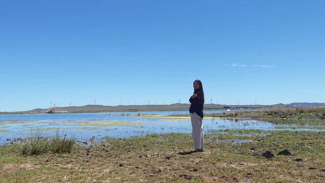 One Asian woman enjoying scenic landscape of salt lake wetland and distant wind turbines in summer in Ulan Hada Volcano Geopark, Ulanqab, Inner Mongolia, China