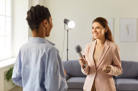 An interviewer smiles as she gives a microphone to female guest during an interview in a studio, with soft lighting and relaxed conversation. Young happy journalist interviewing a woman. 