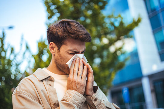 Young Man Sneezing Into Tissue Outdoors in Urban Park During Allergy Season
