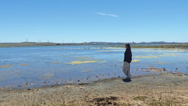 One Asian woman enjoying scenic landscape of salt lake wetland and distant wind turbines in summer in Ulan Hada Volcano Geopark, Ulanqab, Inner Mongolia, China