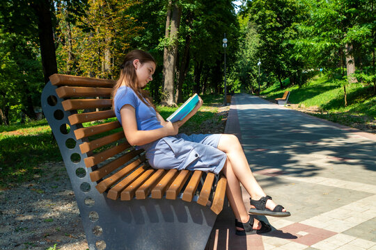 Young woman reading a book on a bench in a sunny city park pathway