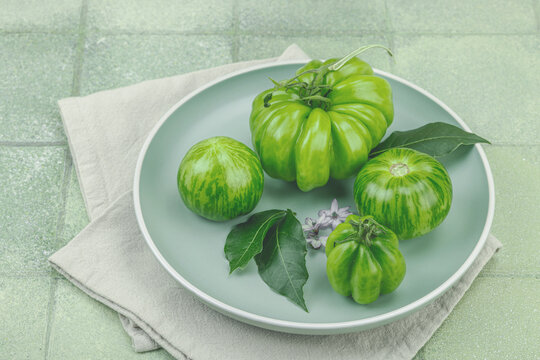Assorted green heirloom tomatoes with leaves and small flowers on a pale plate with a linen napkin