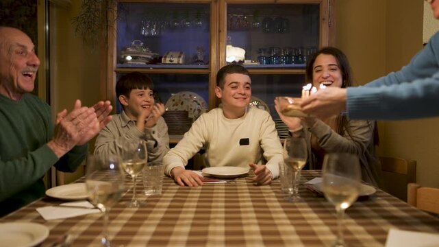 A boy looks up in surprise as a tiramisu cake is placed before him. Family sings happy birthday and claps as he closes his eyes, blows out the candles. Camera pushes in to a close-up. Video 4K.
