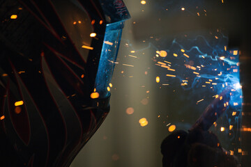 A close-up of a flame-patterned welding helmet with bright sparks glowing in the background, showcasing protective gear used during precision welding inspection in a workshop. © DK_STUDIO