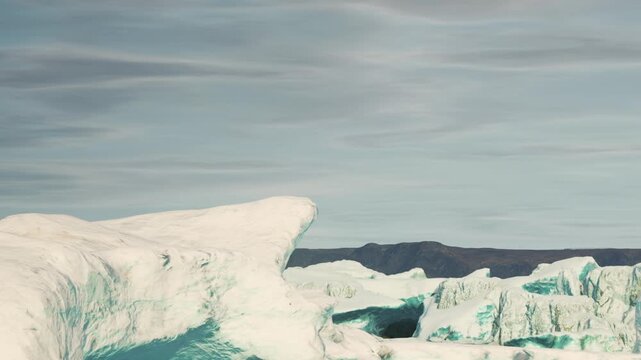 Large icebergs are seen in a remote location with clear skies. The bright blue water contrasts with the white ice. It is cold and the landscape is vast with no signs of human activity.
