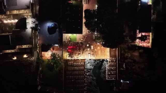 Aerial view of a vibrant bar scene at night along the sea coast, showcasing illuminated outdoor seating, patrons enjoying drinks, and a lively atmosphere with surrounding natural elements. Vacation