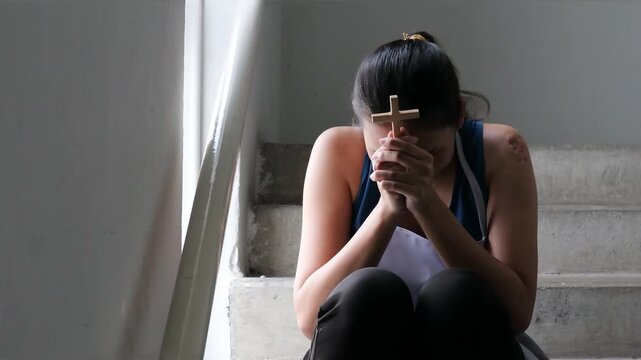 Struggling female worker prays on emergency stairwell, cross in hand, seeking hope during tough times