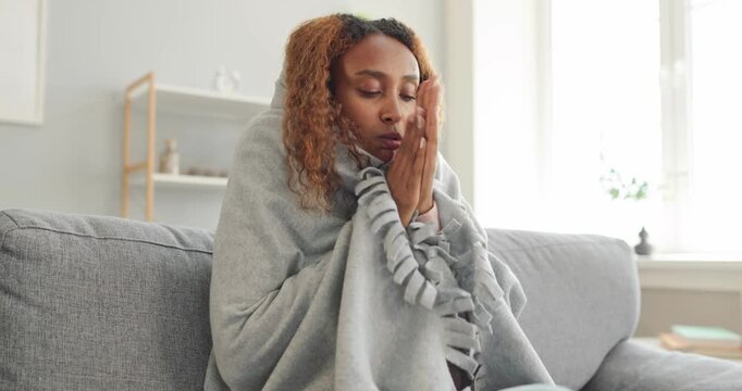 Cold woman shivering under blanket at home. An unwell woman sits on sofa, wrapped in a gray throw beside a bright window in a cozy living room. Winter illness and heating discomfort concept.