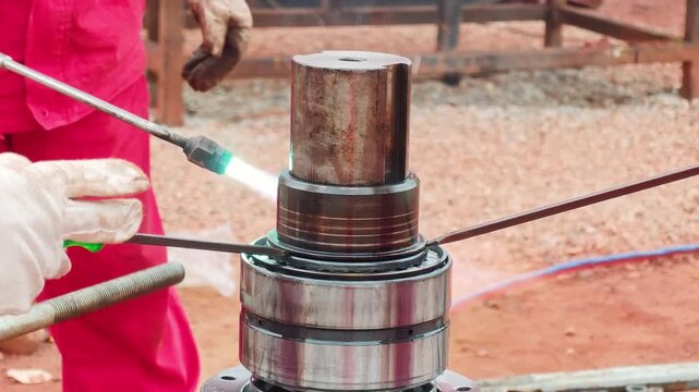 A welder is using a gas blowtorch to apply heat to a steel bearing on a heavy machinery shaft for thermal expansion removal. Industrial working scene, close-up with selective focus.