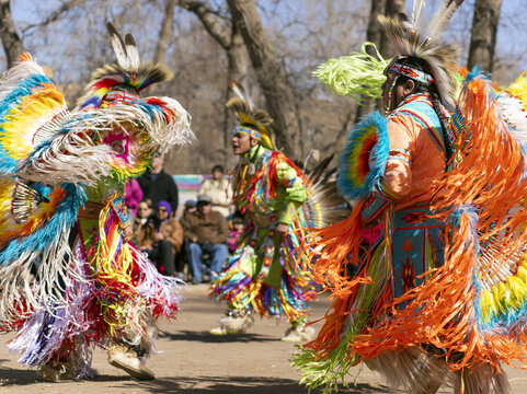Native American dancers in vibrant traditional regalia performing at an outdoor cultural gathering. Indigenous men in colorful fringed clothing and feather headdresses dancing in front of a crowd