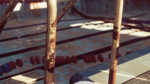 A rusty ladder stands beside an empty swimming pool in a closed facility. Sunlight shines through windows, casting shadows on the tiled floor. The area shows signs of neglect and disuse.