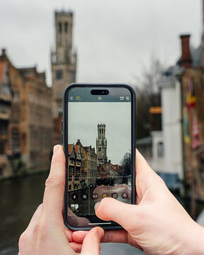 Bruges, Belgium - 03 February 2024: View of the Belfry of Bruges seen through a smartphone camera held by a person, with historic canal-side buildings and a cloudy sky in the background.