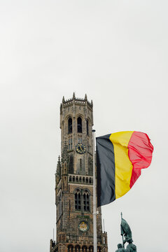 Bruges, Belgium - 03 February 2024: View of the Belfry of Bruges with the Belgian flag waving in the foreground against a cloudy sky.