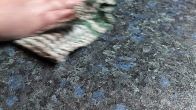 Close-up of a hand using a green and white checkered cloth to wipe and clean a dark, speckled kitchen countertop. Housekeeping chore