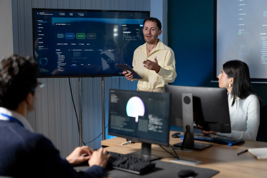 Young male technology consultant presents software dashboard to colleagues during office meeting, using tablet while discussing system data, digital workflow, project planning, and business strategy.