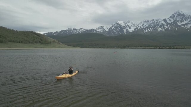 Close up aerial shot of a young man paddling hard in a yellow kayak while a woman row slowly in background on a windy mountain lake, flat log profile