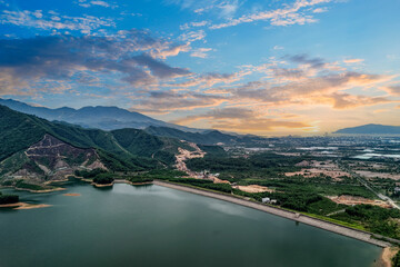 Obraz premium A lake bordered by green areas and mountains in Vietnam. Clouds cover the sky as the sun shines over the water and land