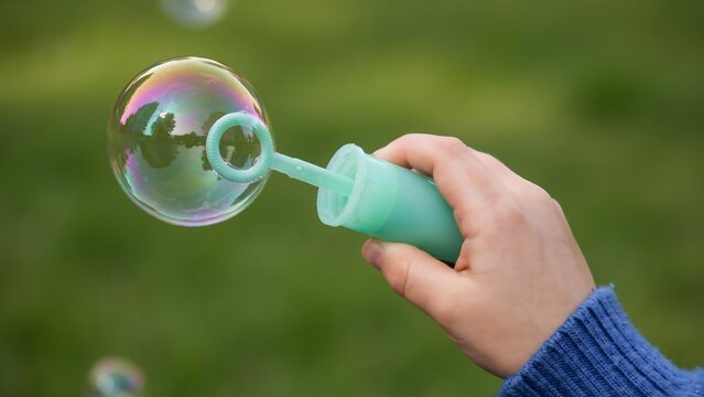 A child hand holding a plastic bubble wand and blowing a large iridescent soap bubble in a green garden