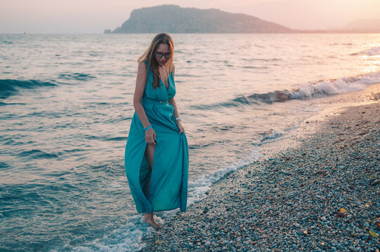 Young woman in a light dress stands by a waterfront railing, enjoying a peaceful sunset over the sea.