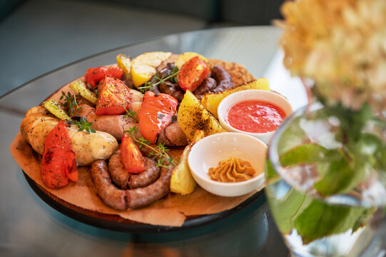 Plate of grilled sausages served with roasted vegetables, herbs, and dipping sauces, placed beside a floral vase on a glass table.