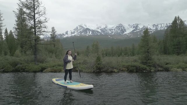 Aerial medium shot of a young woman paddling hard on a sup board during windy weather on a mountain lake, flat log profile