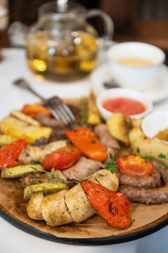 Plate of grilled sausages served with roasted vegetables, herbs, and dipping sauces, placed beside a floral vase on a glass table.