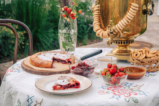 A golden samovar with bagels, tea cups, homemade pie, and jam on a white tablecloth set for tea in a garden.