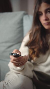 Close-up of a young Hispanic woman's hand using a TV remote while sitting on her sofa, representing home entertainment, media choice, technology, and modern lifestyle.
