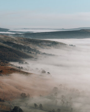 View of Mam Tor rising above a thick blanket of morning mist covering the valley floor with rolling hills and scattered trees in Hope Valley, England, United Kingdom.