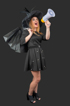 Woman in a black dress and hat holding shopping bags and a megaphone, loudly announcing sales, discounts and promotions with expressive open mouthed call in studio retail setting