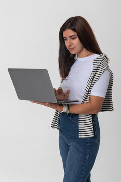 Young woman standing in a white studio, holding a slim laptop and typing while looking at the screen, portraying modern remote work, study and digital connection lifestyle