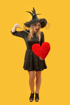 Young woman in witch costume with black hat and dress, casting a magic spell with her wand over a fluffy red heart on a bright yellow background, whimsical romance feel